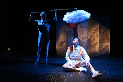 Szenenfoto „Heart Shaped Box” von wonderlendt performances. Eine Performer*in in einem weißen Kleid ist auf dem dunklen Bühnenboden. Eine Performer*in in einem schwarzen Ganzkörperanzug sthet im Hintergrund und hält der sitzenden Performer*in an einer Stange eine große weiße flauschige Wolken über den Kopf.