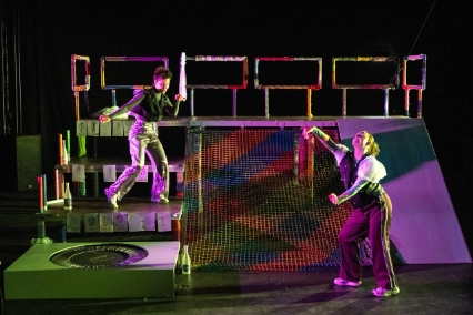 Szenenfoto „Der Urspung der Welt” von frau emma gelb. Zwei Performer*innen tanzen ausdrucksstark auf der Bühne. Auf der Bühne steht eine Art Gerüst links mit Stufen, rechts einer Art Rutschplatte und in der Mitte einem bunten Netz. Eine Performer*in steht auf einer der Stufen, die andere Person tanzt vor der Rutschplatte.