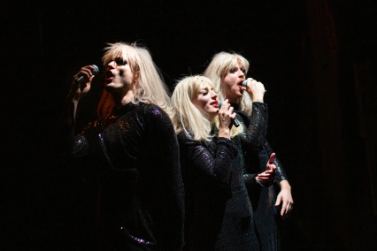 Szenenfoto „How to survive with HILDEGARD KNEF. Eine Rettung in zehn Liedern” von Tommy Wiesner und Ensemble – Drei Performer*innen mit langen blonden Perücken, roten Lippen und schwarzen Paillettenkleidern stehen jeweils mit einem Mikrofon singend nebeneinanderstehend