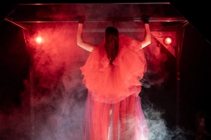 Szenenfoto „REDSHIFT – Tales of gainig power” von tilted collective – Zu sehen ist eine Person in einem bauschigen Tüllkleid, die eingehüllt in Bühnenleben Klimmzüge an einer Metallstange macht. Scheinwerfer werfen rotes Licht auf die Person.