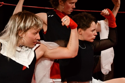 Szenenfoto „fetzen” von kollektiv fetzen – Nahaufnahme von drei Performer*innen, die auf dem Boden hocken und mit wütendem Blick nach rechts ihre Armmuskeln zeigen. Alle drei sind in Schwarz, Rot und Weiß gekleidet. Zwie tragen schwarze Boxershorts auf deren Bund Fetzen steht.