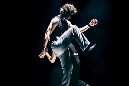 Szenenfoto „Motion in Sound„ von Kollektiv mis – zwei Performer*innen schreiten in einem hellen Scheinwerferkegel umeinander – die hinter Person gebückt und kleiner, die vordere Person macht gerade einen großen Schritt und hat ein Knie sehr weit nach oben gezogen.