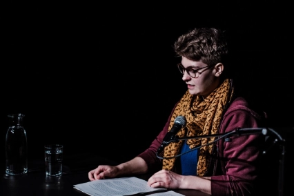 Szenenfoto „etwas mitgenommen” von Mia Wiebke Thies – eine Performer*in an einem Tisch sitzen und liest von einem Manuskript.