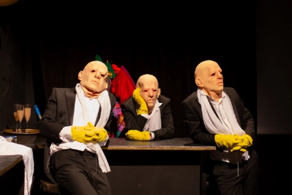 Szenenfoto „MEPHISTO. UNDER PRESSURE.” von Theater an der Glocksee – Drei Personen sind an einem Tisch. All drei sind identisch gekleidet und tragen die gleichen Gummimasken über dem kompletten Kopf.