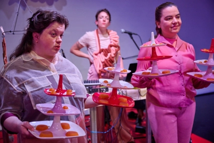 Szenenfoto „Tod in der Tupperbox” von Frl. Wunder AG & Theater Thikwa – Zwei Performer*innen präsentieren Essen auf bunten Etageren. Im Hintergrund steht eine Performer*in. Statt einer Schürze hat sie einen Stoffdarm umgehängt.