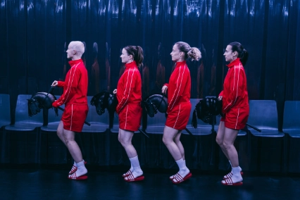 Szenenfoto „HYPER” von cindy+cate. Vier Performer*innen reiten auf Steckenpferden von rechts nach links über die dunkle Bühne. Alle vier tragen das gleiche Outfit: kurze rote Hosen, rote Jacken, weiße Sportsocken und rot-weiß-gestreifte Badelatschen der Marke Adidas.