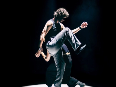 Szenenfoto „Motion in Sound„ von Kollektiv mis – zwei Performer*innen schreiten in einem hellen Scheinwerferkegel umeinander – die hinter Person gebückt und kleiner, die vordere Person macht gerade einen großen Schritt und hat ein Knie sehr weit nach oben gezogen.