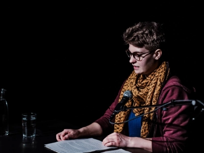 Szenenfoto „etwas mitgenommen” von Mia Wiebke Thies – eine Performer*in an einem Tisch sitzen und liest von einem Manuskript.