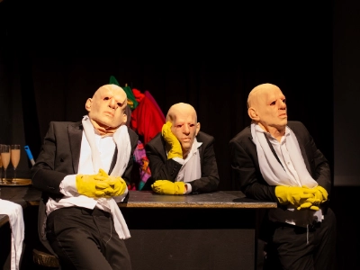 Szenenfoto „MEPHISTO. UNDER PRESSURE.” von Theater an der Glocksee – Drei Personen sind an einem Tisch. All drei sind identisch gekleidet und tragen die gleichen Gummimasken über dem kompletten Kopf.