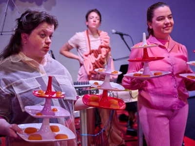 Szenenfoto „Tod in der Tupperbox” von Frl. Wunder AG & Theater Thikwa – Zwei Performer*innen präsentieren Essen auf bunten Etageren. Im Hintergrund steht eine Performer*in. Statt einer Schürze hat sie einen Stoffdarm umgehängt.