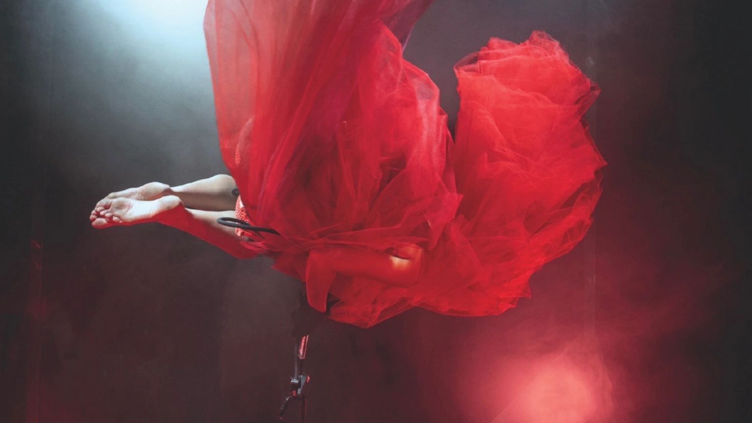 Szenenfoto REDSHIFT – Tales of gainig power von tilted collective – Zu sehen ist eine Person in einem bauschigen roten Tüllkleid, die in der Luft zu schweben scheint, sich dafür aber an einem Stahlseil festhält. Vom Körper sind lediglich die nackten Unterschenkel und Füße zu sehen und ein Arm. Ein Scheinwerfer wirft rotes Licht auf die Person.