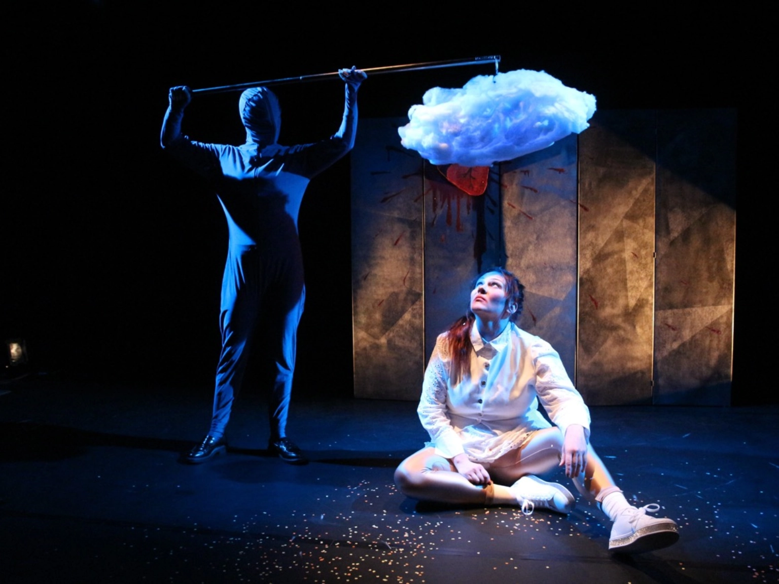 Szenenfoto „Heart Shaped Box” von wonderlendt performances. Eine Performer*in in einem weißen Kleid ist auf dem dunklen Bühnenboden. Eine Performer*in in einem schwarzen Ganzkörperanzug sthet im Hintergrund und hält der sitzenden Performer*in an einer Stange eine große weiße flauschige Wolken über den Kopf.