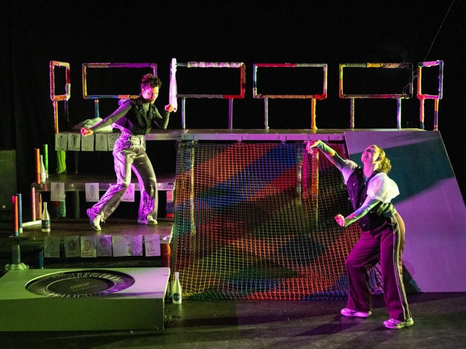 Szenenfoto „Der Urspung der Welt” von frau emma gelb. Zwei Performer*innen tanzen ausdrucksstark auf der Bühne. Auf der Bühne steht eine Art Gerüst links mit Stufen, rechts einer Art Rutschplatte und in der Mitte einem bunten Netz. Eine Performer*in steht auf einer der Stufen, die andere Person tanzt vor der Rutschplatte.