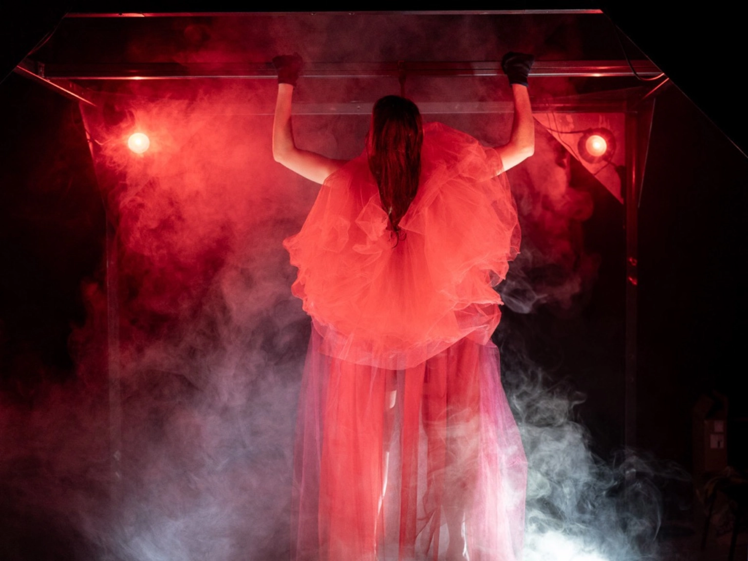 Szenenfoto „REDSHIFT – Tales of gainig power” von tilted collective – Zu sehen ist eine Person in einem bauschigen Tüllkleid, die eingehüllt in Bühnenleben Klimmzüge an einer Metallstange macht. Scheinwerfer werfen rotes Licht auf die Person.