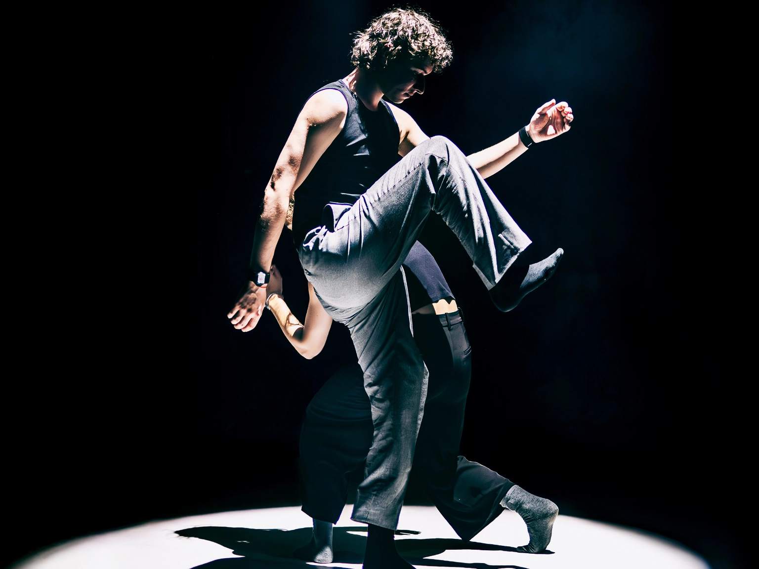 Szenenfoto Motion in Sound von Kollektiv mis – zwei Performer*innen schreiten in einem hellen Scheinwerferkegel umeinander – die hinter Person gebückt und kleiner, die vordere Person macht gerade einen großen Schritt und hat ein Knie sehr weit nach oben gezogen.