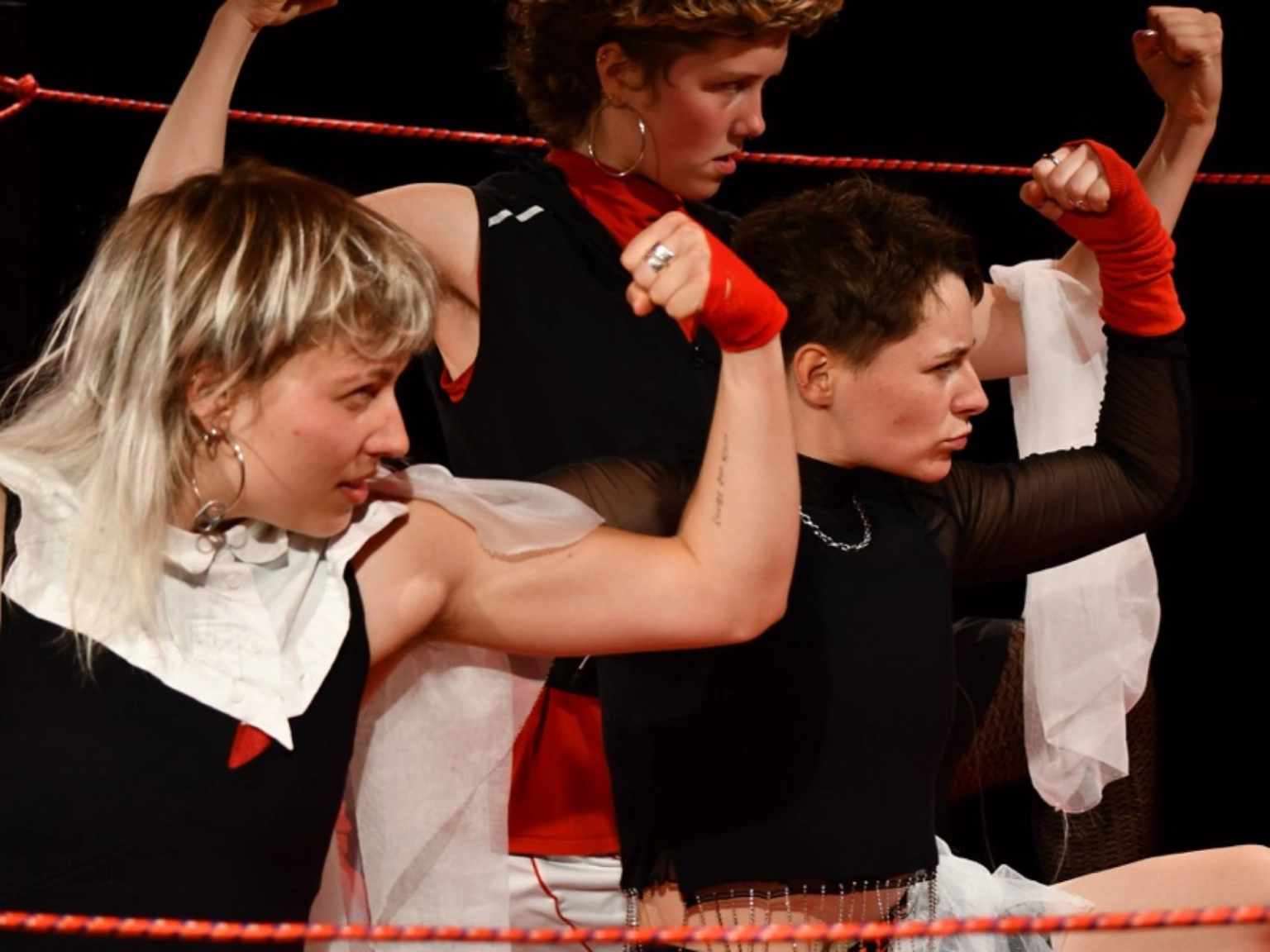 Szenenfoto „fetzen” von kollektiv fetzen – Nahaufnahme von drei Performer*innen, die auf dem Boden hocken und mit wütendem Blick nach rechts ihre Armmuskeln zeigen. Alle drei sind in Schwarz, Rot und Weiß gekleidet. Zwie tragen schwarze Boxershorts auf deren Bund Fetzen steht.