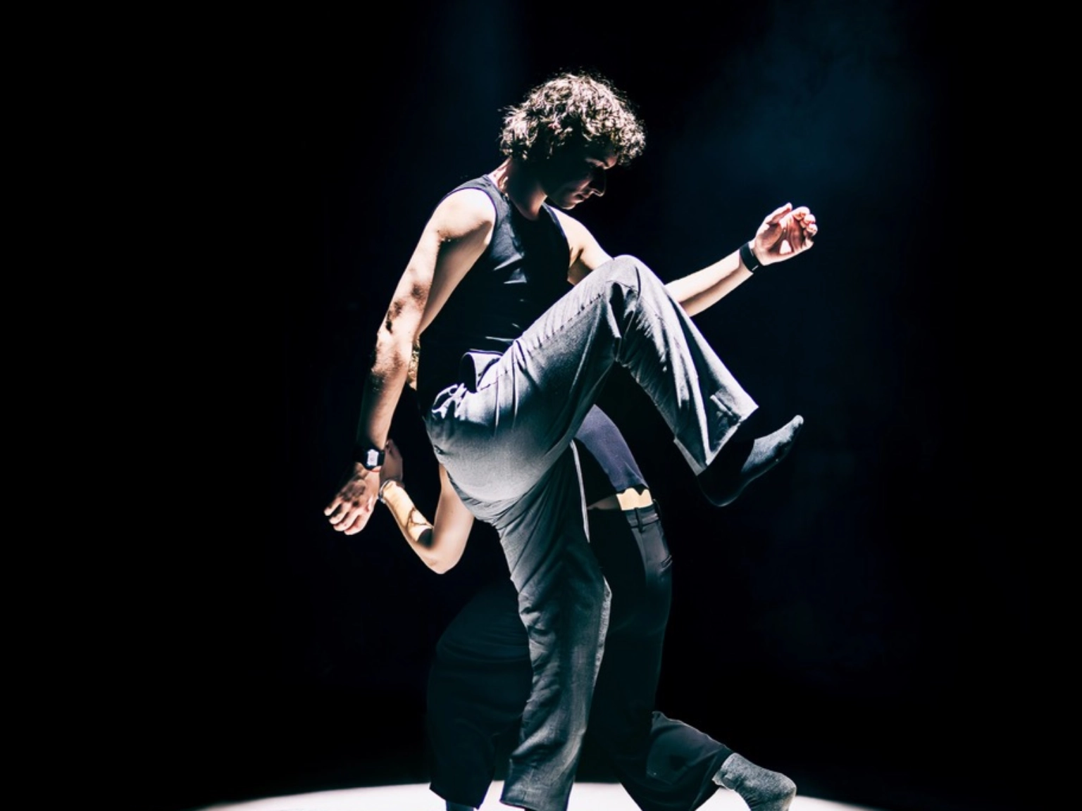 Szenenfoto „Motion in Sound„ von Kollektiv mis – zwei Performer*innen schreiten in einem hellen Scheinwerferkegel umeinander – die hinter Person gebückt und kleiner, die vordere Person macht gerade einen großen Schritt und hat ein Knie sehr weit nach oben gezogen.