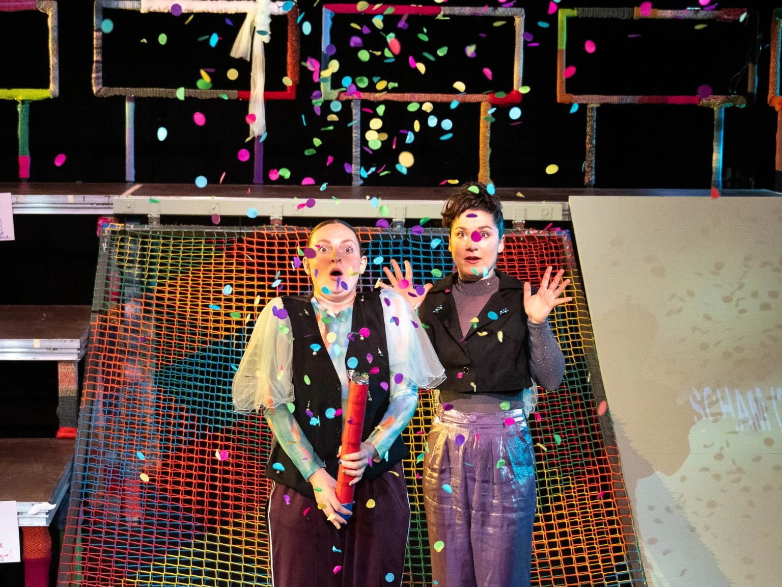 Szenenfoto Der Urspung der Welt von frau emma gelb. Zwei Performer*innen stehen auf der Bühne. Hinter ihnen steht eine Art Gerüst mit links Stufen, rechts einer Art Rutschplatte und in der Mitte einem bunten Netz. Die Performer*innen stehen davor. Eine Person hat gerade eine Konfettikanone ausgelöst. Bunte Konfetti regnet auf sie herab. Beide schauen überrascht, erschrocken, freudig.