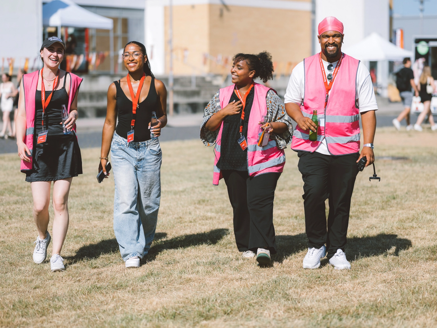 Impression eines Einsatzes des Awarenesskollektivs WirWollen. Vier Personen gehen über eine trockene Wiese, im Hintergrund sind Bühnen und Pavillons zu sehen, drei der vier Personen tragen eine pinke Weste.