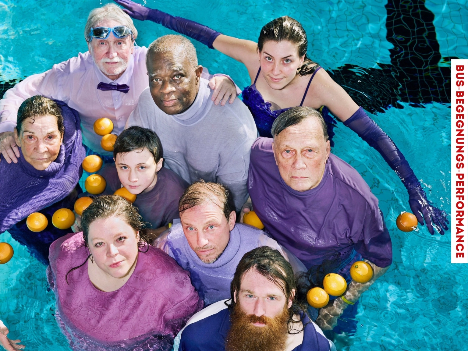 Neun Menschen stehen in Lilafarbenen Outfits in einem Schwimmbecken. Das Foto wird von oben gemacht