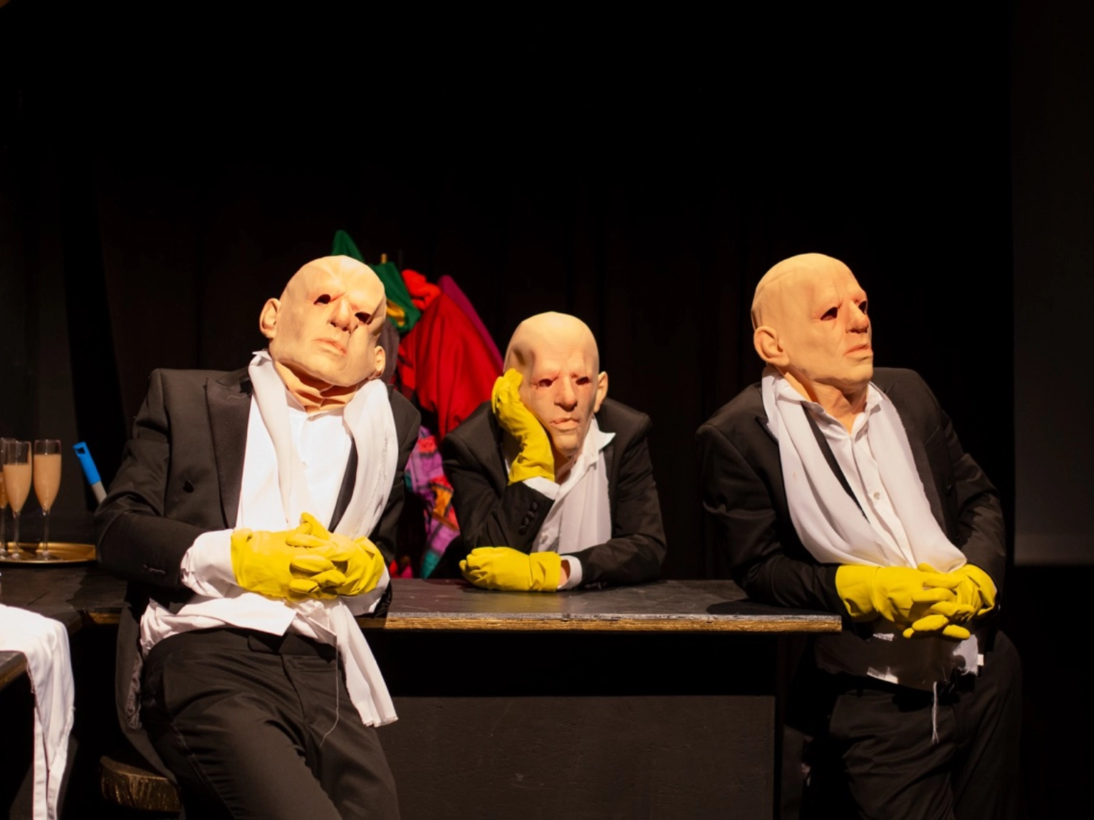 Szenenfoto „MEPHISTO. UNDER PRESSURE.” von Theater an der Glocksee – Drei Personen sind an einem Tisch. All drei sind identisch gekleidet und tragen die gleichen Gummimasken über dem kompletten Kopf.