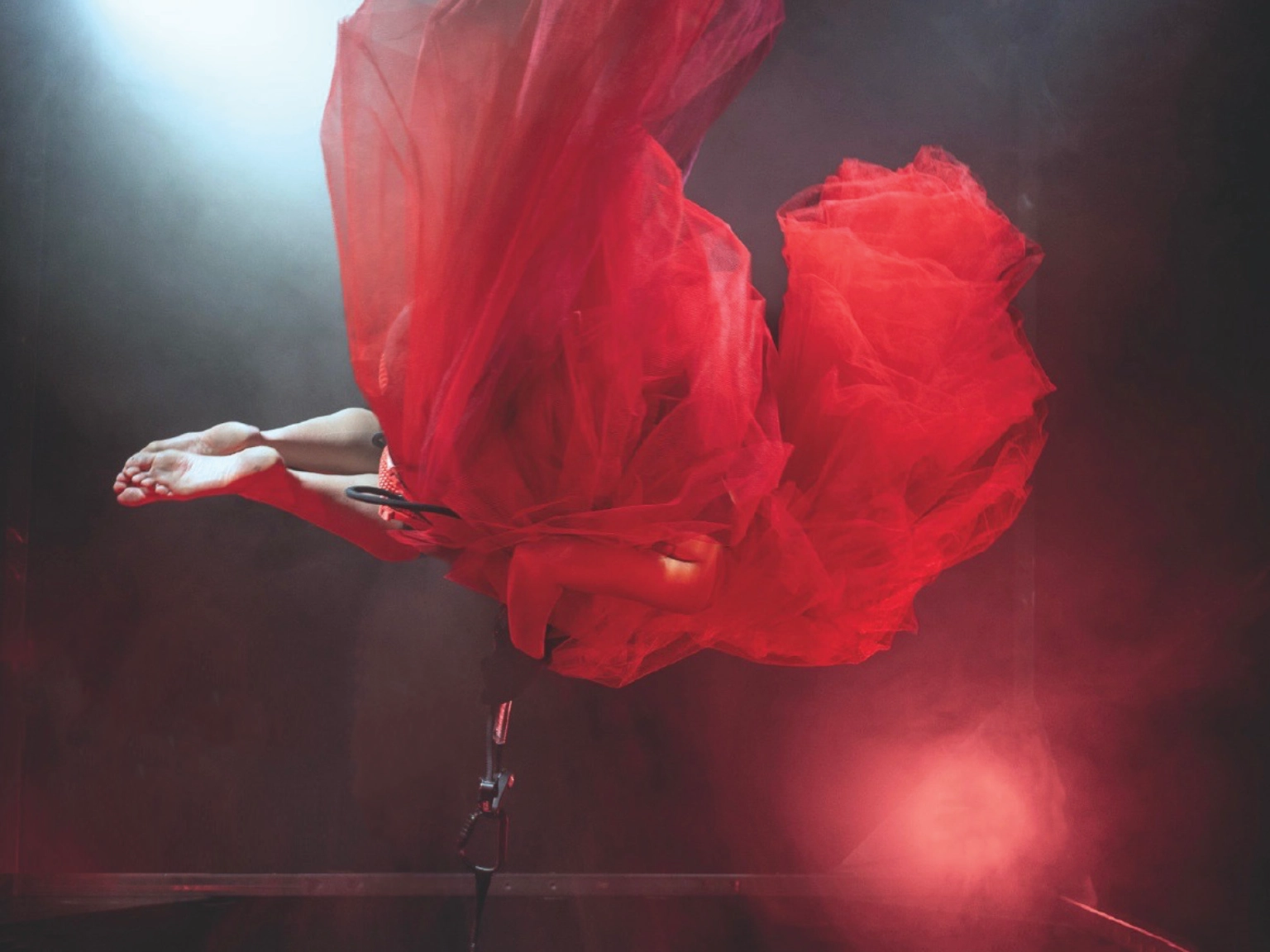 Szenenfoto REDSHIFT – Tales of gainig power von tilted collective – Zu sehen ist eine Person in einem bauschigen roten Tüllkleid, die in der Luft zu schweben scheint, sich dafür aber an einem Stahlseil festhält. Vom Körper sind lediglich die nackten Unterschenkel und Füße zu sehen und ein Arm. Ein Scheinwerfer wirft rotes Licht auf die Person.