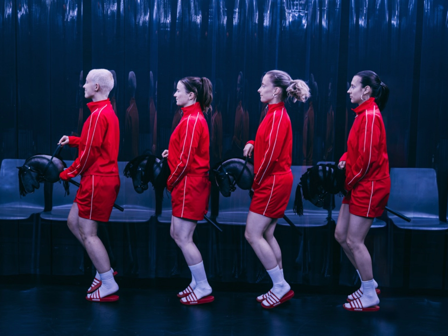 Szenenfoto „HYPER” von cindy+cate. Vier Performer*innen reiten auf Steckenpferden von rechts nach links über die dunkle Bühne. Alle vier tragen das gleiche Outfit: kurze rote Hosen, rote Jacken, weiße Sportsocken und rot-weiß-gestreifte Badelatschen der Marke Adidas.