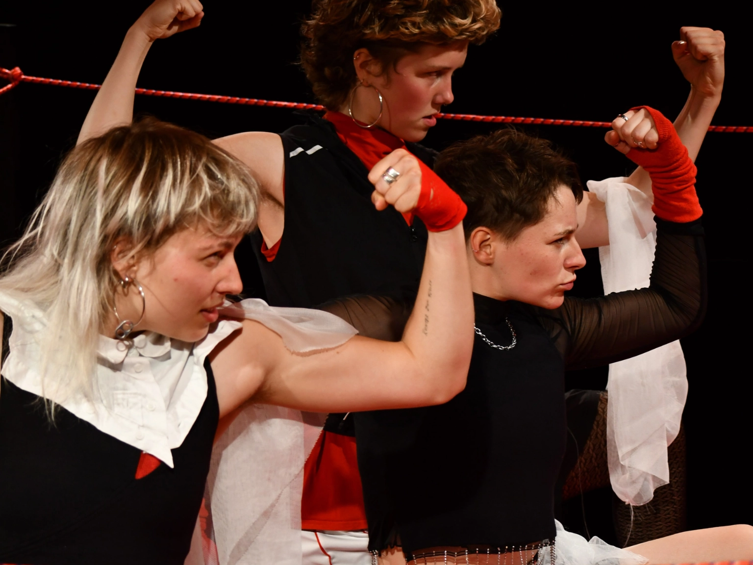 Szenenfoto Fetzen von Kollektiv fetzen – Nahaufnahme von drei Performer*innen, die auf dem Boden hocken und mit wütendem Blick nach rechts ihre Armmuskeln zeigen. Alle drei sind in Schwarz, Rot und Weiß gekleidet. Zwie tragen schwarze Boxershorts auf deren Bund Fetzen steht.