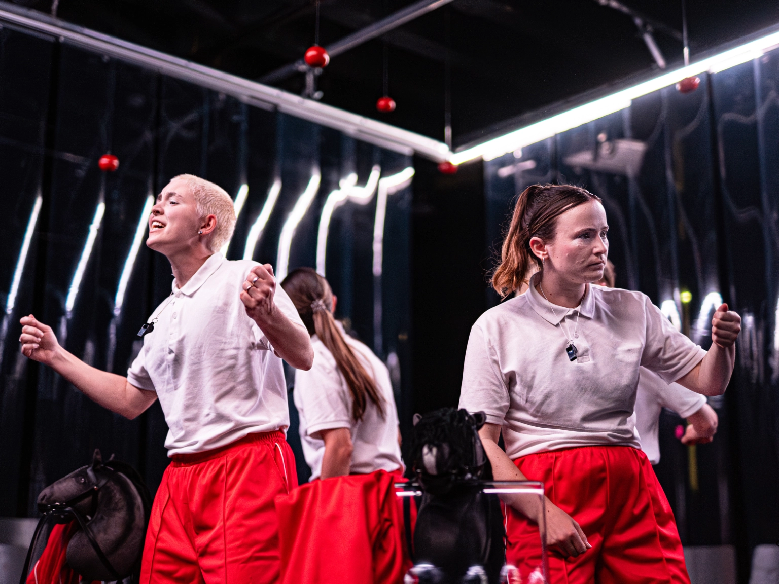 Szenenfoto HYPER von cindy+cate. Vier Performer*innen stehen mit dem Rücken zueinander in einem Kreis. Alle vier tragen das gleiche Outfit: Eine rote Hose und eine weißes T-Shirt. Zwei Performer*innen sieht man von vorne. Sie sind in einem intensiven Spiel mit starker Mimik und Gesten. 