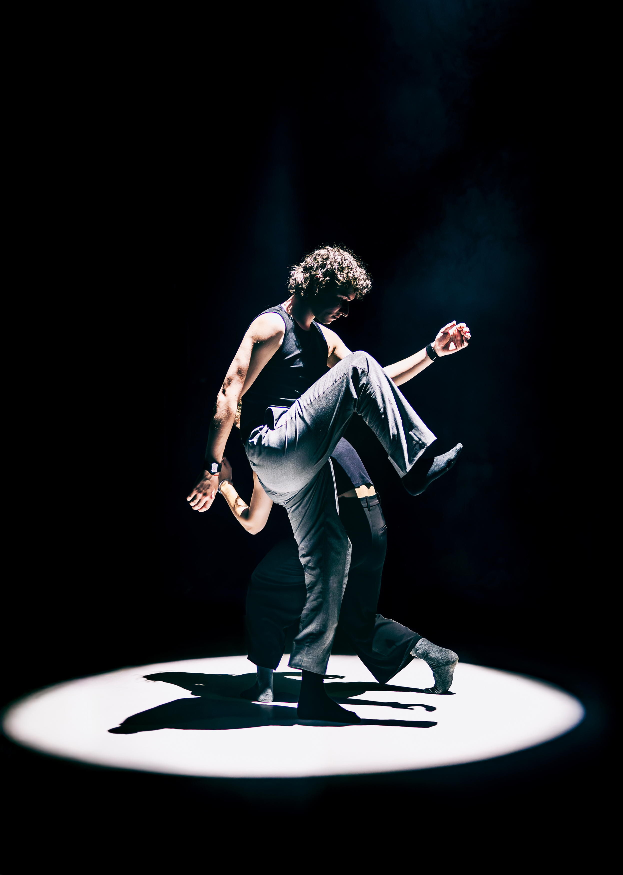 Szenenfoto Motion in Sound von Kollektiv mis – zwei Performer*innen schreiten in einem hellen Scheinwerferkegel umeinander – die hinter Person gebückt und kleiner, die vordere Person macht gerade einen großen Schritt und hat ein Knie sehr weit nach oben gezogen.