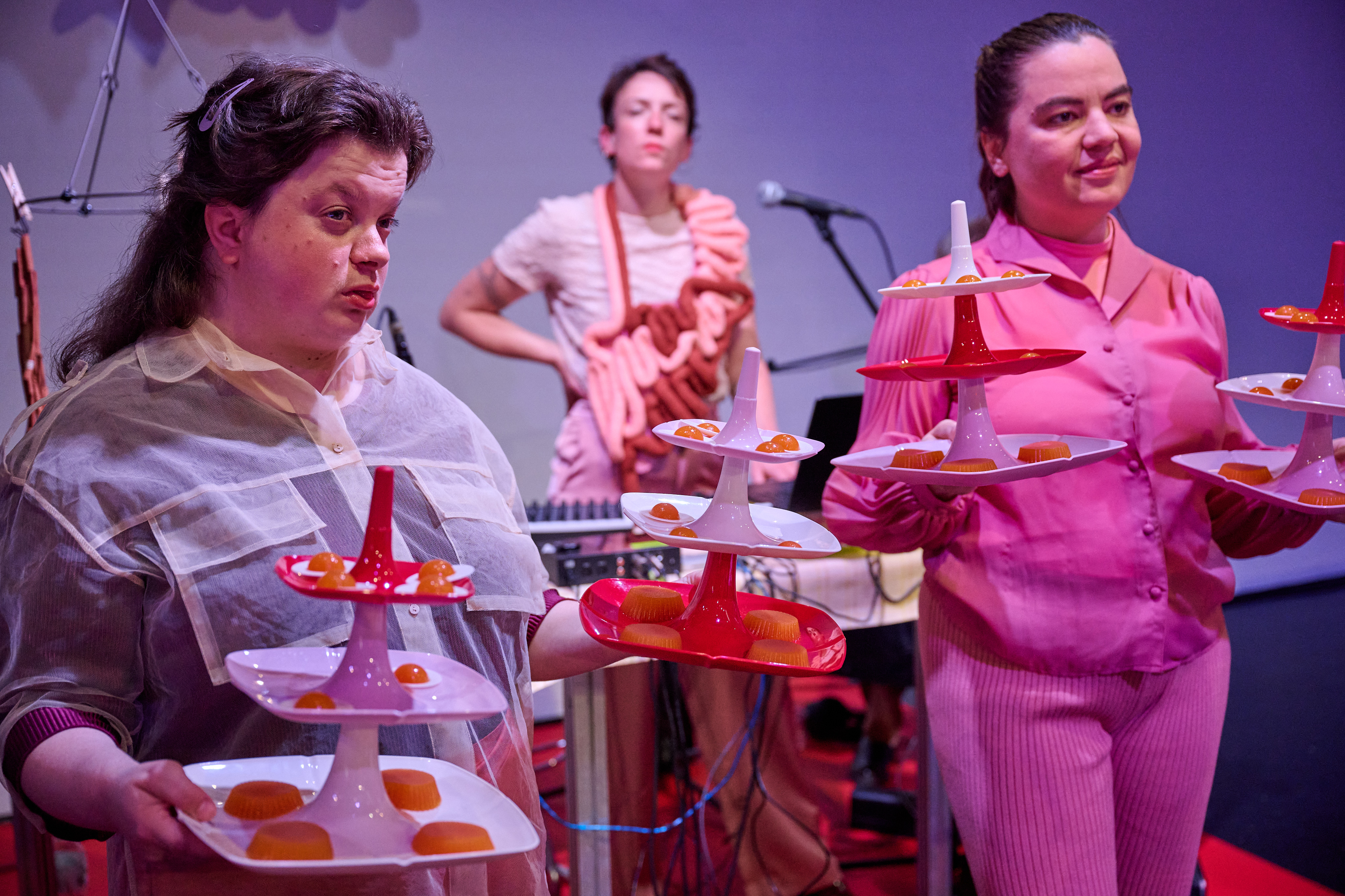 Szenenfoto Tod in der Tupperbox von Frl. Wunder AG gemeinsam mit Theater Thikwa – Zwei Performer*innen präsentieren Essen auf bunten Etageren. Im Hintergrund steht eine Performer*in. Statt einer Schürze hat sie einen Stoffdarm umgehängt.