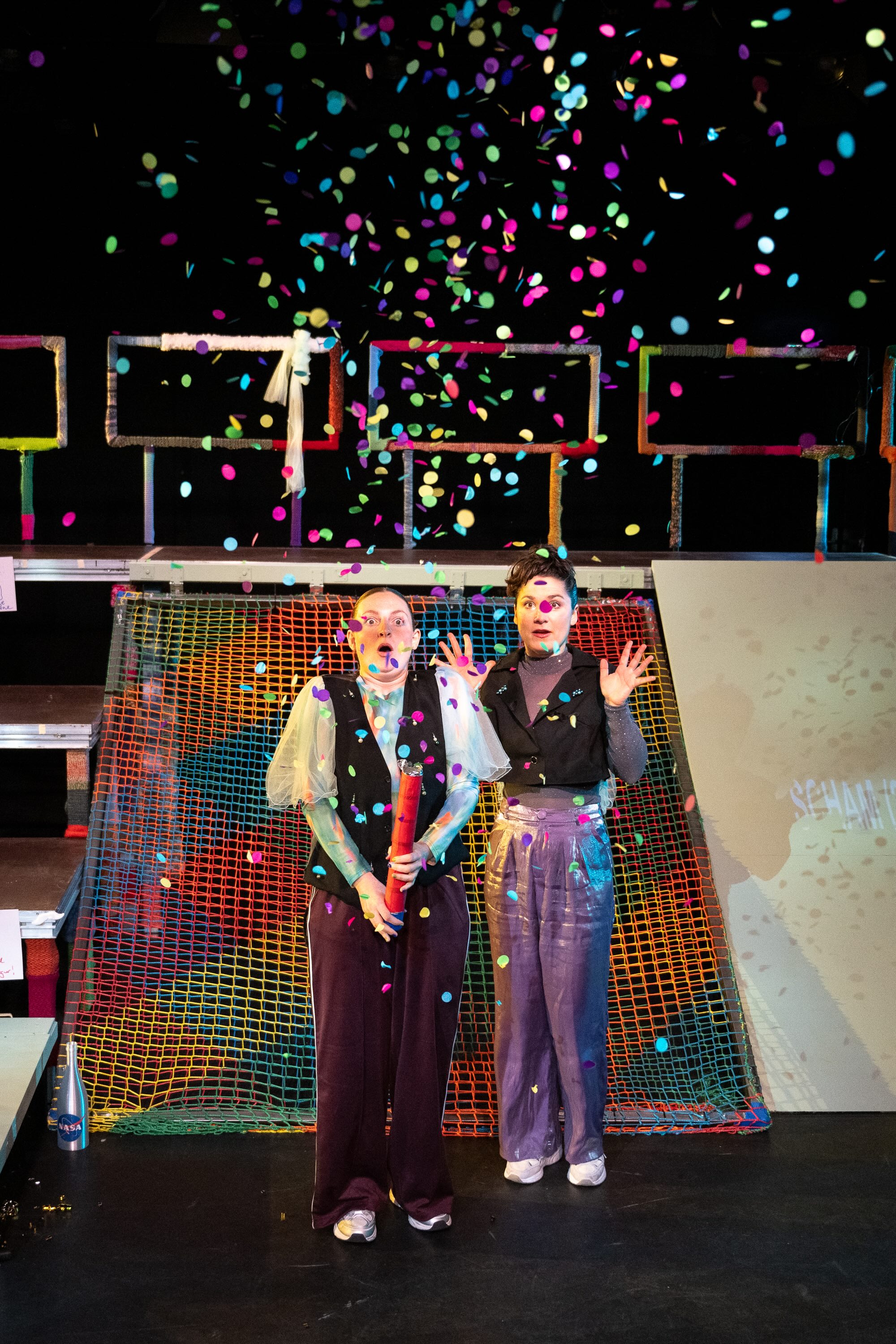 Szenenfoto Der Urspung der Welt von frau emma gelb. Zwei Performer*innen stehen auf der Bühne. Hinter ihnen steht eine Art Gerüst mit links Stufen, rechts einer Art Rutschplatte und in der Mitte einem bunten Netz. Die Performer*innen stehen davor. Eine Person hat gerade eine Konfettikanone ausgelöst. Bunte Konfetti regnet auf sie herab. Beide schauen überrascht, erschrocken, freudig.