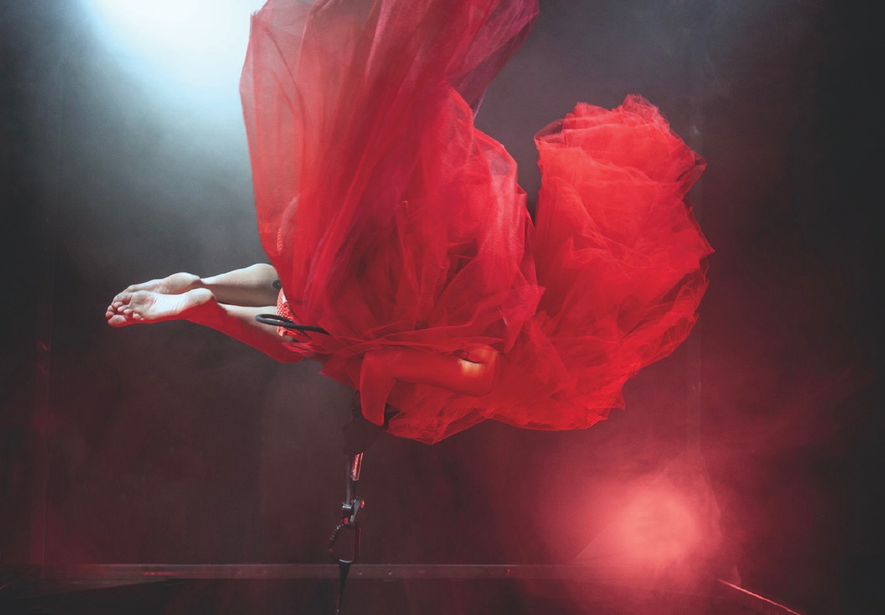 Szenenfoto REDSHIFT – Tales of gainig power von tilted collective – Zu sehen ist eine Person in einem bauschigen roten Tüllkleid, die in der Luft zu schweben scheint, sich dafür aber an einem Stahlseil festhält. Vom Körper sind lediglich die nackten Unterschenkel und Füße zu sehen und ein Arm. Ein Scheinwerfer wirft rotes Licht auf die Person.